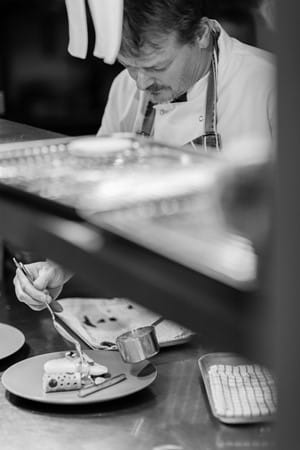 Black-and-white photo of Sean Kelly, Head Chef at Station Road, leaning over the pass in his chef whites, carefully plating a dish with a small utensil in hand